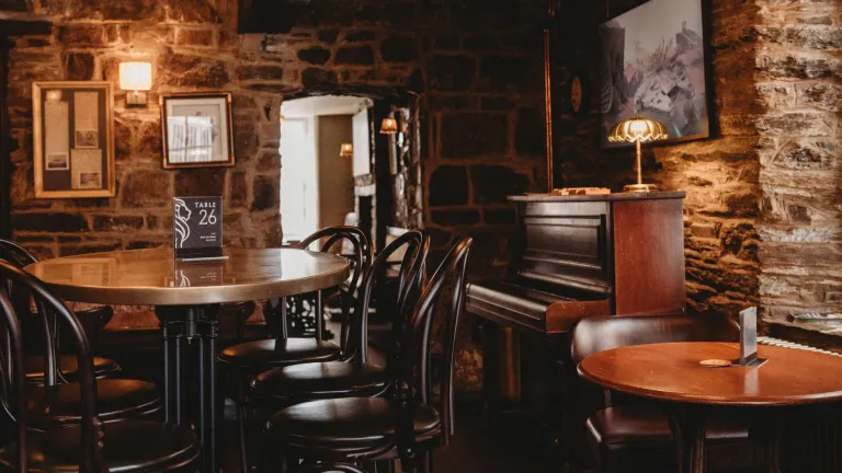 Tables, chairs and a piano in a stone walled lounge area of a hotel.