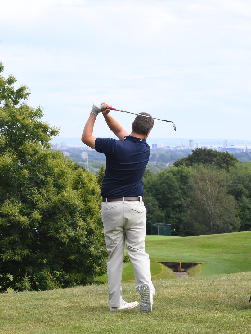 Golfer having taken a swing next to his golf bag and fellow player with view of Cardiff beyond.