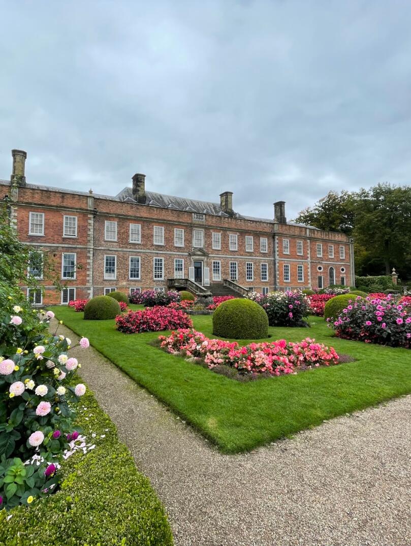 A grand house with beautiful flowers and round shrubbery in the forecourt.