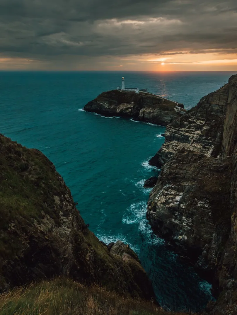 Person standing on a coast path overlooking a lighthouse at sunset.