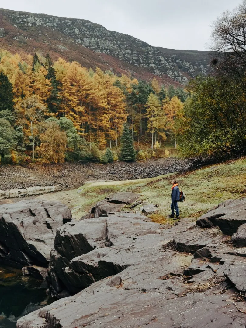 A walker by a lake surronded by rocks and an autumnal forest.