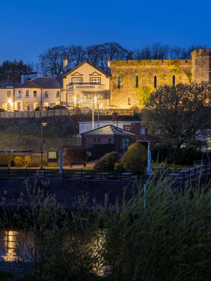 External shot of a castle hotel lit up at night, with a river running beside it.