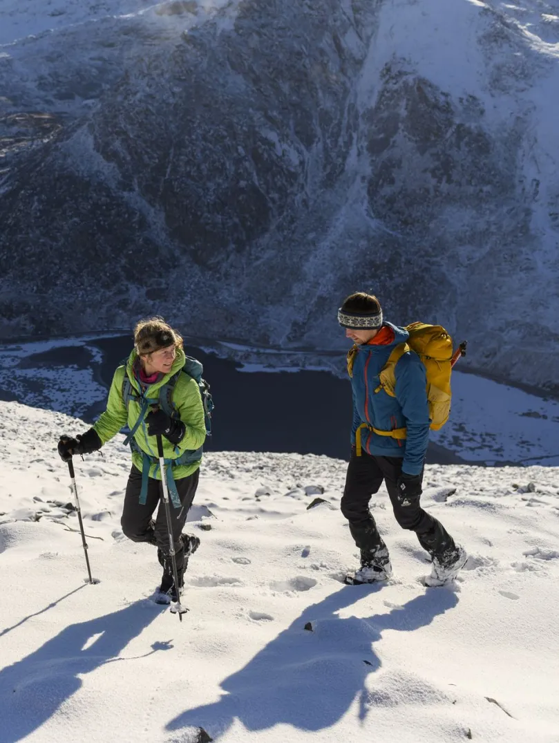 A couple walking on a snowy mountain with a frozen lake below.