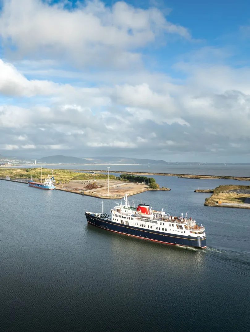 A cruise ship heading toward a port With mountains and the shoreline in the background.