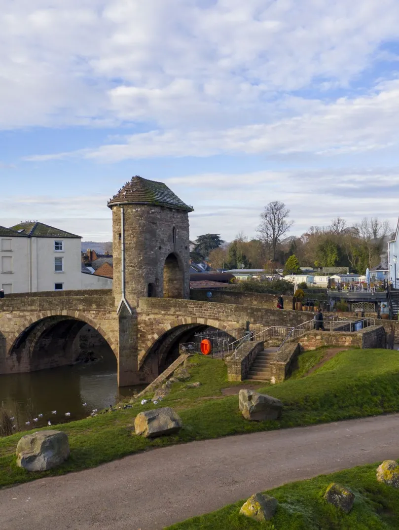 View of stone bridge and gatehouse with town buildings and a riverside path under a partly cloudy sky.