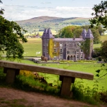 A large manor house with its towers covered in creeping ivy viewed from a seating bench.