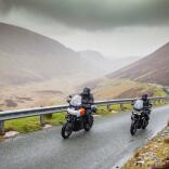 Two motorcyclists driving on a road amongst mountains.
