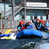 A group kitted out in helmets and lifej ackets using their oars to take on the rapids in a blue dinghy.