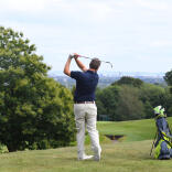 Golfer having taken a swing next to his golf bag and fellow player with view of Cardiff beyond.