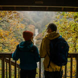 A couple on a viewing platform looking at a waterfall surrounded by autumnal trees.
