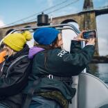 Group of ladies on a speed boat with one taking a photo of a bridge.