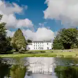 A white manor surrounded by trees reflecting in a lake.