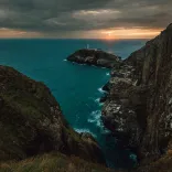 Person standing on a coast path overlooking a lighthouse at sunset.