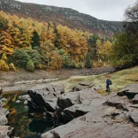 A walker by a lake surronded by rocks and an autumnal forest.