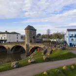 View of stone bridge and gatehouse with town buildings and a riverside path under a partly cloudy sky.