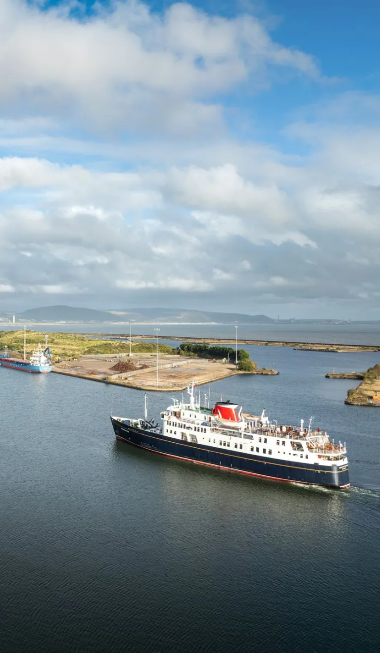 A cruise ship heading toward a port With mountains and the shoreline in the background.
